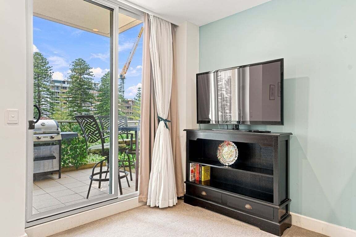 Una habitación con puerta corrediza de vidrio que da a un balcón, un televisor sobre un mueble de madera oscura, y una parrilla afuera.
