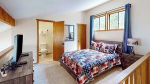 Un dormitorio con cama, escritorio, televisor, ventana con cortinas y puerta que da a un baño.