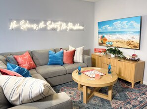 Ein Wohnzimmer mit grauem Sofa, bunten Kissen, einem Holztisch, einem Scrabble-Spiel und einem Neon-Schild.