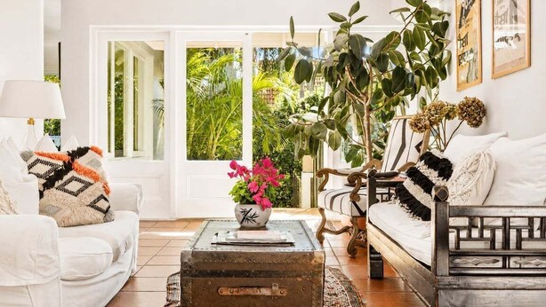 Uma sala de estar com um sofá branco, um banco de madeira, um baú antigo usado como mesa de centro e uma grande janela com vista para o verde.