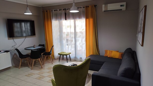 A living room with a sofa, a dining table with chairs, a TV mounted on the wall, and an air conditioning unit. - HUGE BALCONY APARTMENT NEXT TO ONE SALONIKA OUTLET (Thessaloniki)