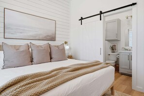 Un dormitorio con cama, almohadas, un edredón, una mesita de noche, una lámpara fijada a la pared y una puerta que da al baño.