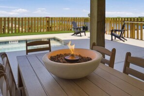 Una mesa de madera con un cuenco que contiene fuego encendido, sillas exteriores, una piscina y una valla de madera.