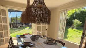 A dining area with a glass table, dark wooden chairs, a woven pendant light, and a view of a garden through large windows.