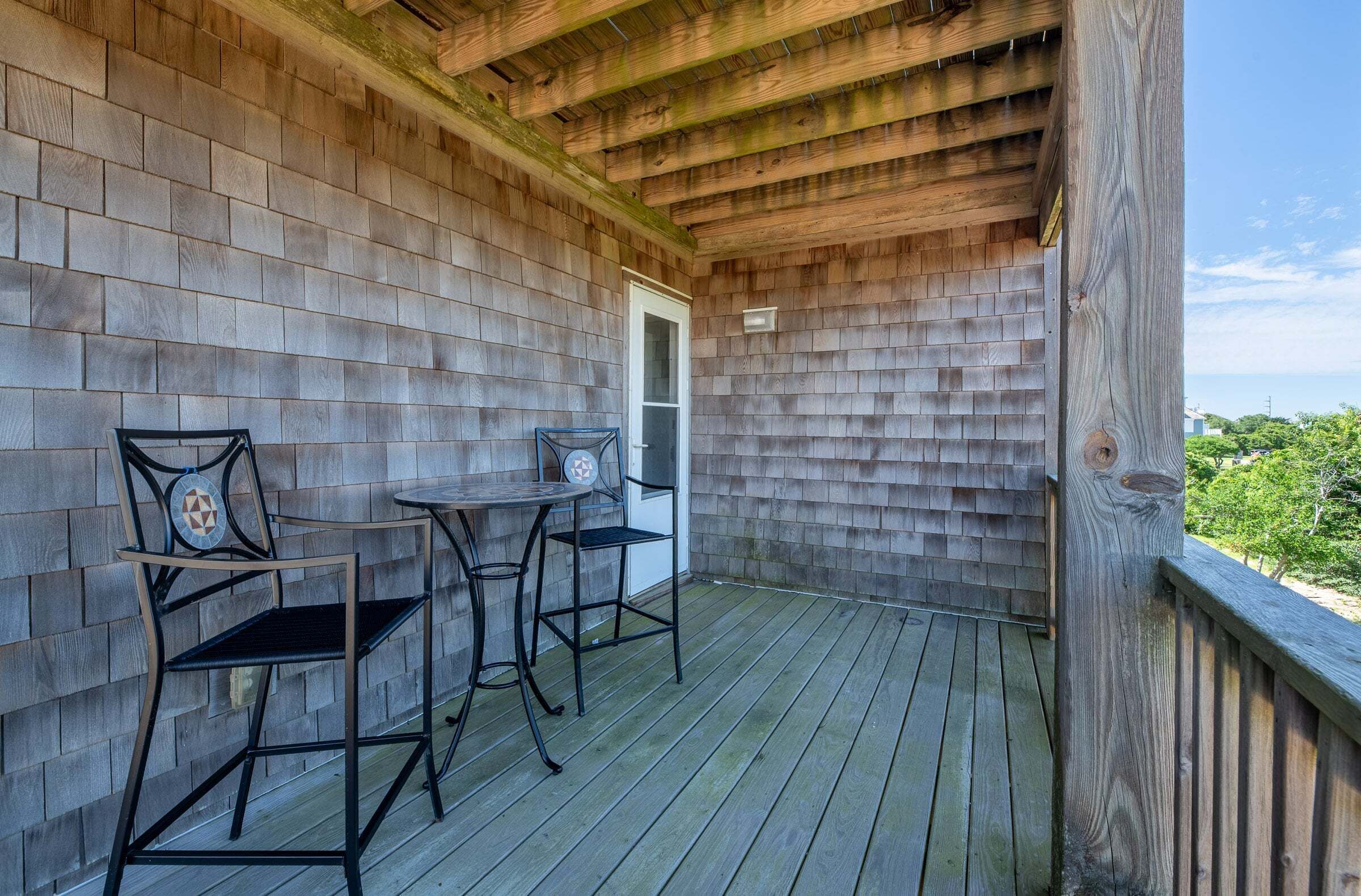 A wooden deck with two metal chairs and a small table, a wooden door, and a view of greenery.
