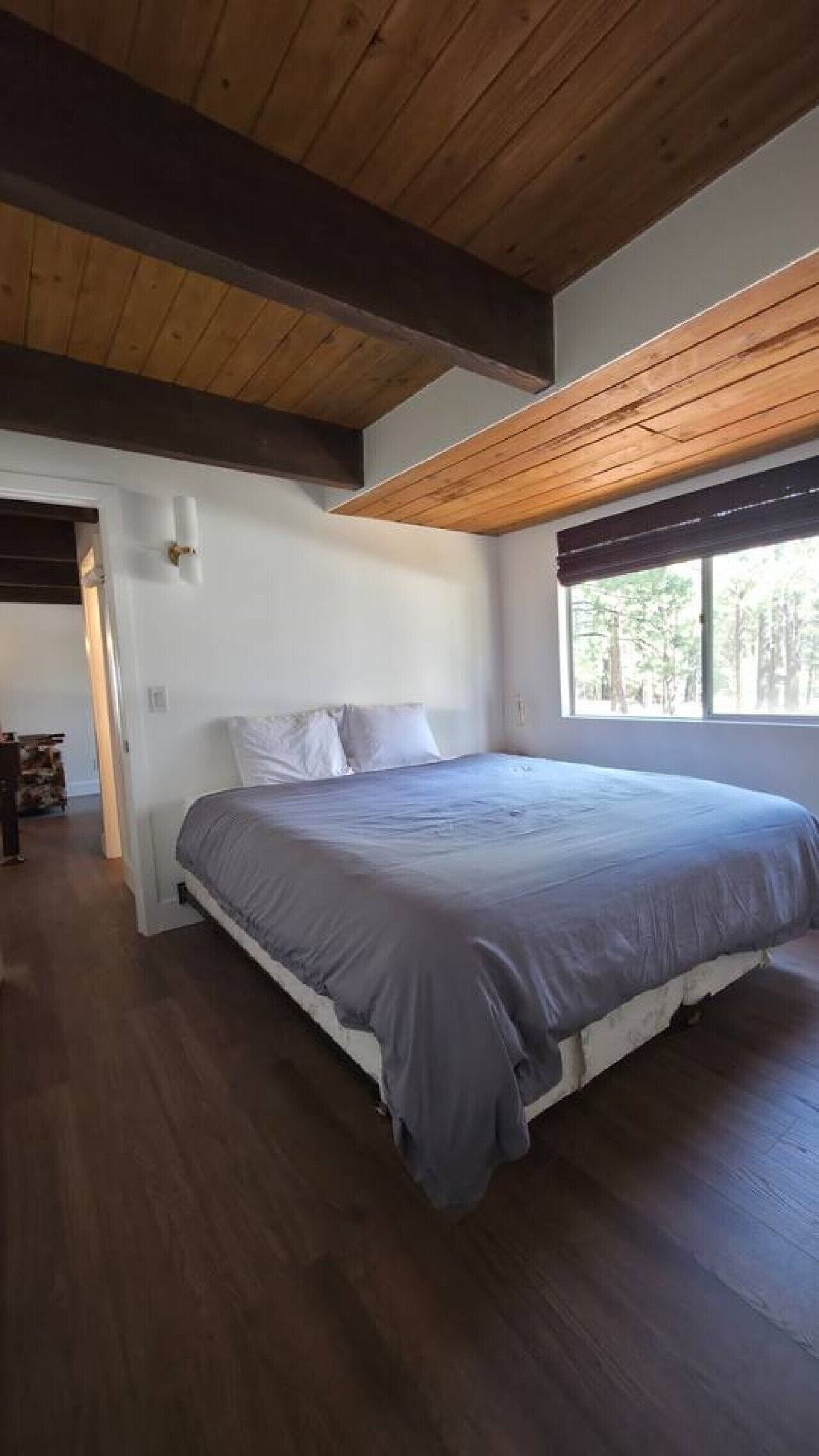 Un dormitorio con una cama grande, vigas de madera en el techo, una ventana con vista a árboles y una puerta que da a otra habitación.