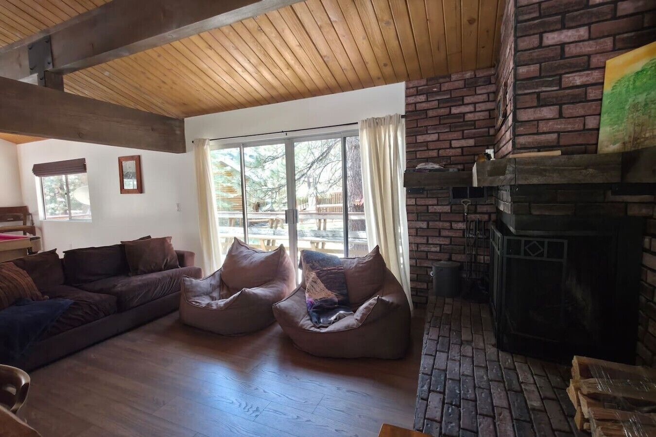 Una sala de estar acogedora con techo de madera, chimenea de ladrillo, ventana grande con cortinas y un sofá con sillones de peluche.
