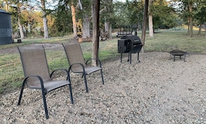 Two outdoor chairs, a grill, and a fire pit in a wooded area.