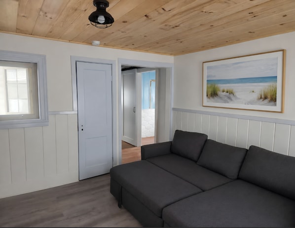 Une pièce avec un canapé gris, un plafond en bois, une porte blanche et un tableau représentant une plage.