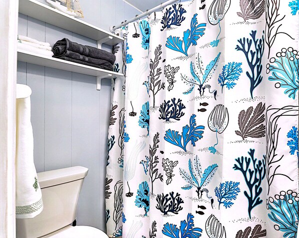 Une salle de bain avec des toilettes, une étagère avec des serviettes pliées et un rideau de douche orné d’un motif de feuilles bleues et noires.