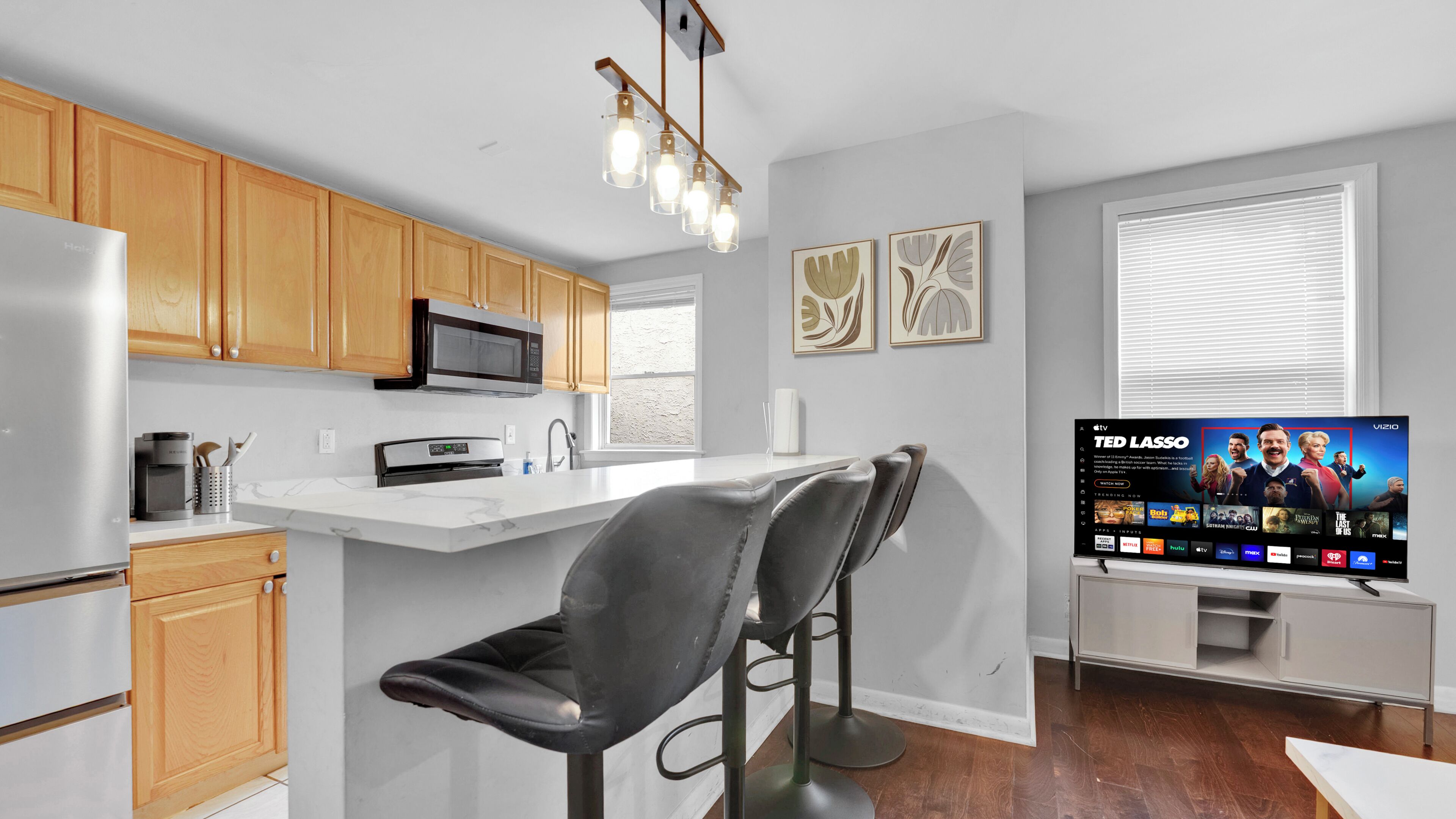 A modern kitchen with wooden cabinets, a stainless steel refrigerator, a microwave, a built-in oven, a bar stool, and a flat-screen TV displaying a TV show.