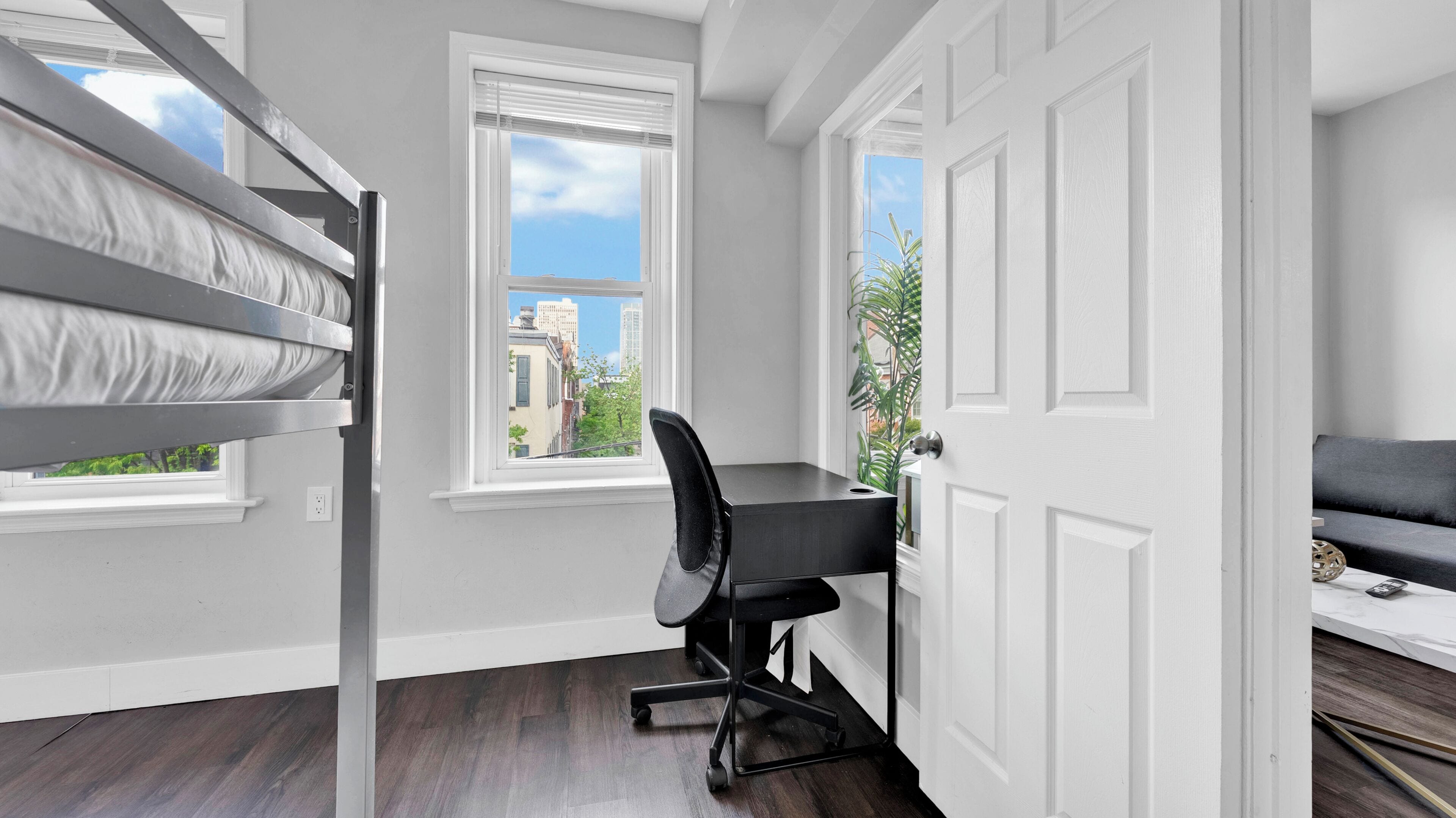A room with a bunk bed, a desk with a chair, a sofa, and a view of buildings through the windows.