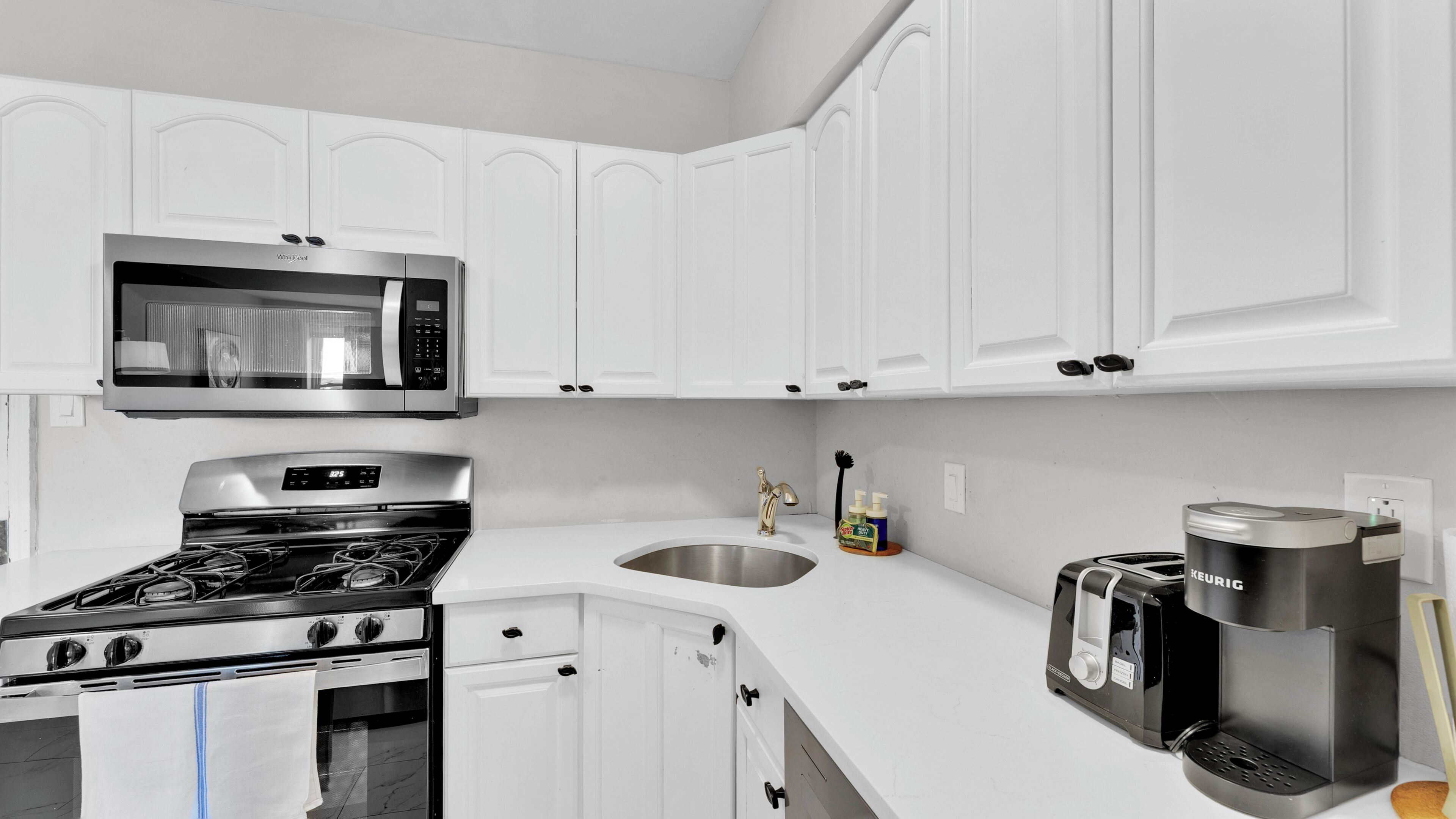 A kitchen with white cabinets, a microwave, a stove, a sink, and a coffee maker.