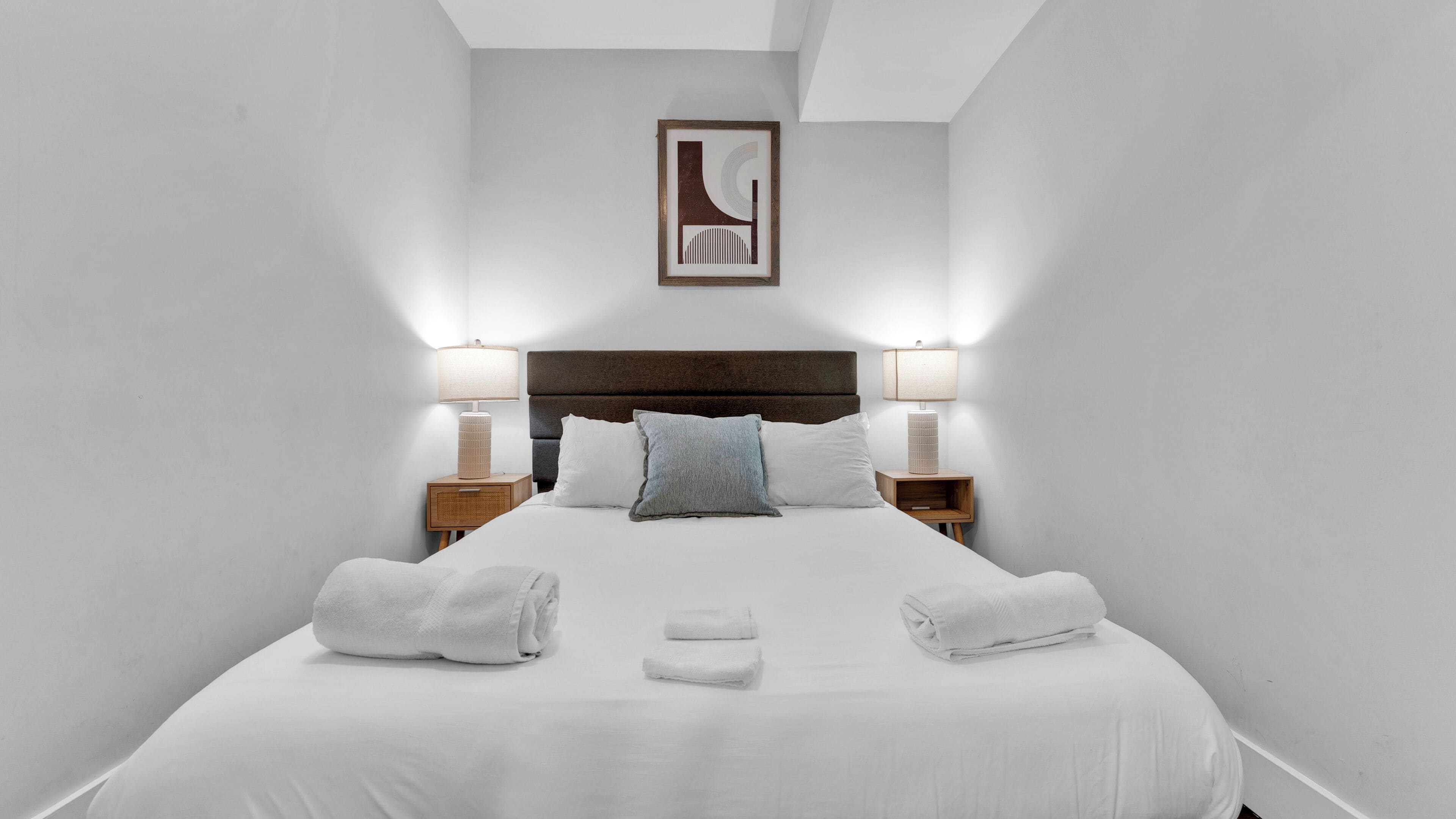 A hotel room with a large bed, two bedside tables with lamps, a headboard, and a framed picture on the wall.