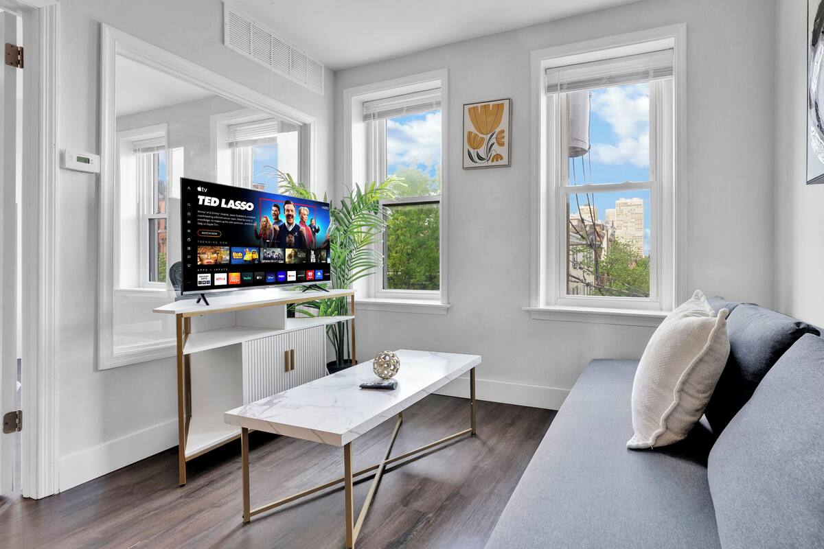 A modern living room with a flat-screen TV, a white coffee table, a grey sofa, and large windows with a city view.