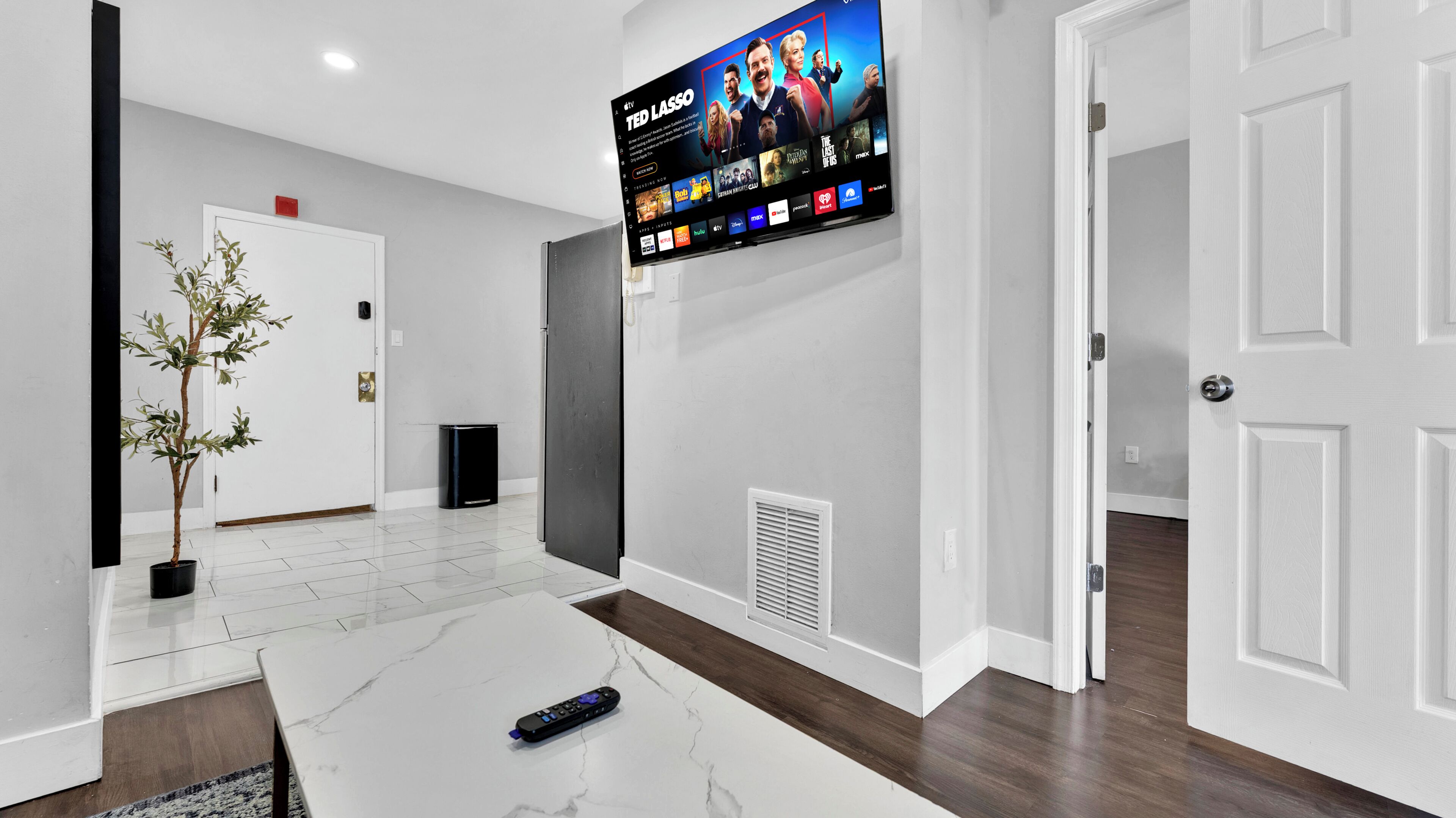 A modern living room with a flat-screen TV displaying a movie, a potted plant, a remote control, and a marble coffee table.