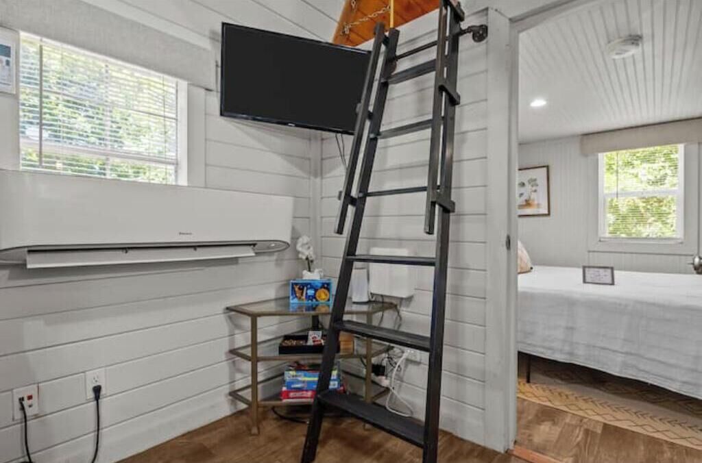 A room with a bed, a TV mounted on the wall, a ladder, and a shelf with items.