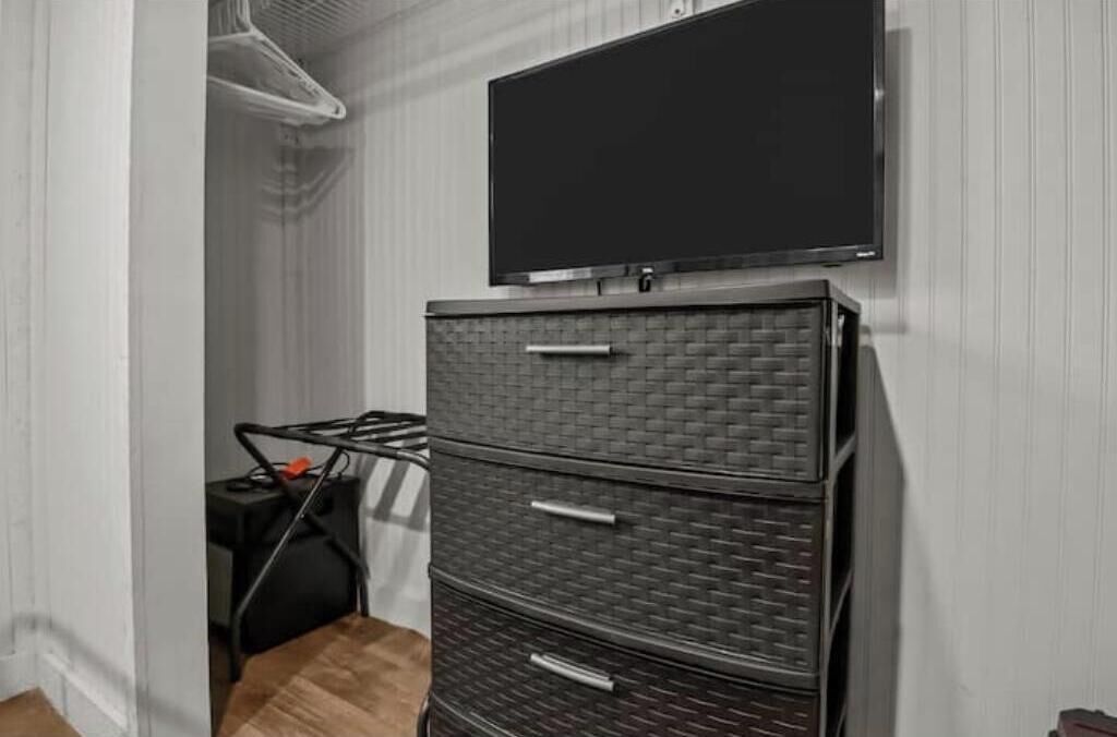 A room with a flat-screen TV mounted on the wall, a wicker storage unit with drawers, and a small table with a chair.