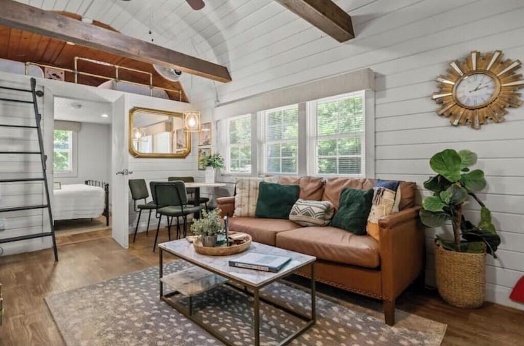 A cozy living room with a brown leather sofa, a coffee table, and a large mirror. There is a ladder in the corner and a window with a view of trees.