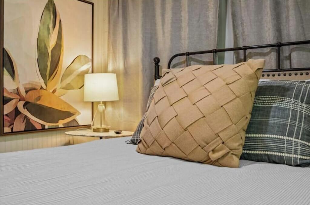 A bed with a woven pillow, a bedside lamp, and a framed artwork on the wall.