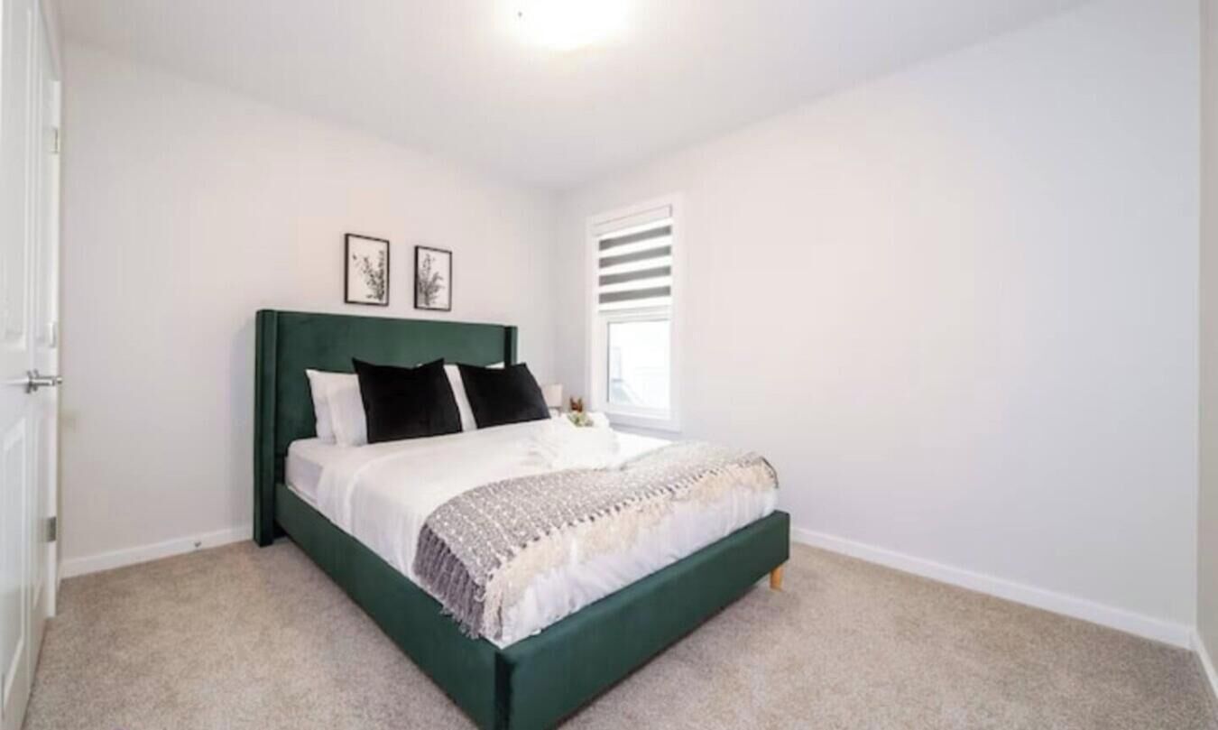 Un dormitorio con cama, paredes blancas, una ventana con persianas y dos cuadros en la pared.