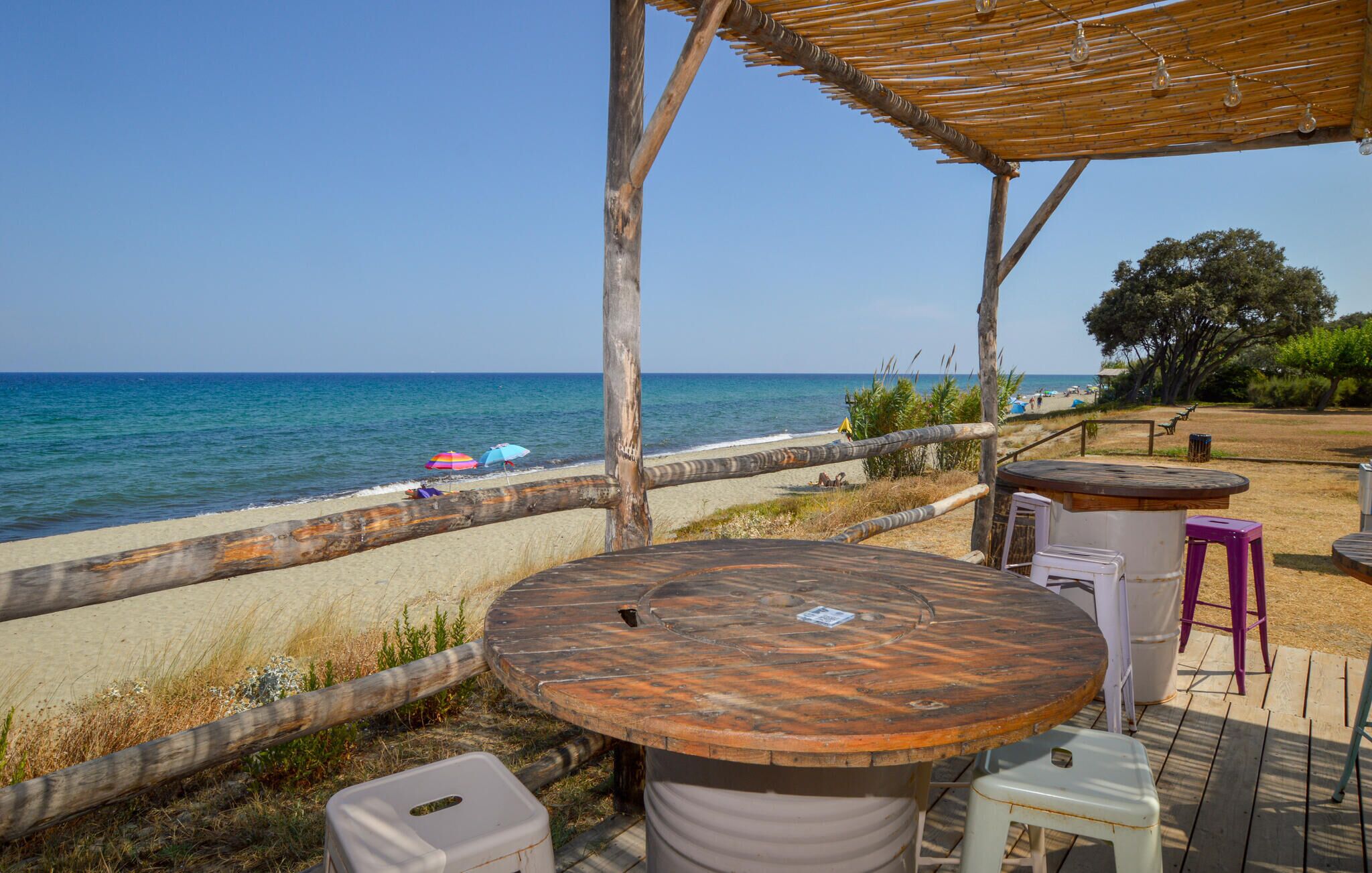 Eine Holzterrasse mit einem runden Tisch und Hockern, mit Blick auf einen Strand mit Sonnenschirmen und freiem Meerblick.