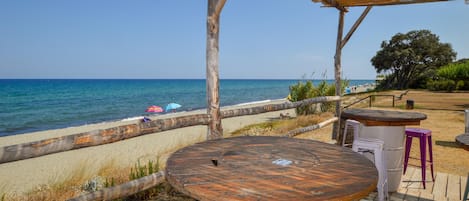 A wooden deck with a round table and stools, overlooking a beach with umbrellas and a clear view of the sea.