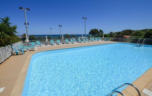 Ein klares, blaues Schwimmbecken mit Liegestühlen und Sonnenschirmen, umgeben von einem Zaun und Palmen.