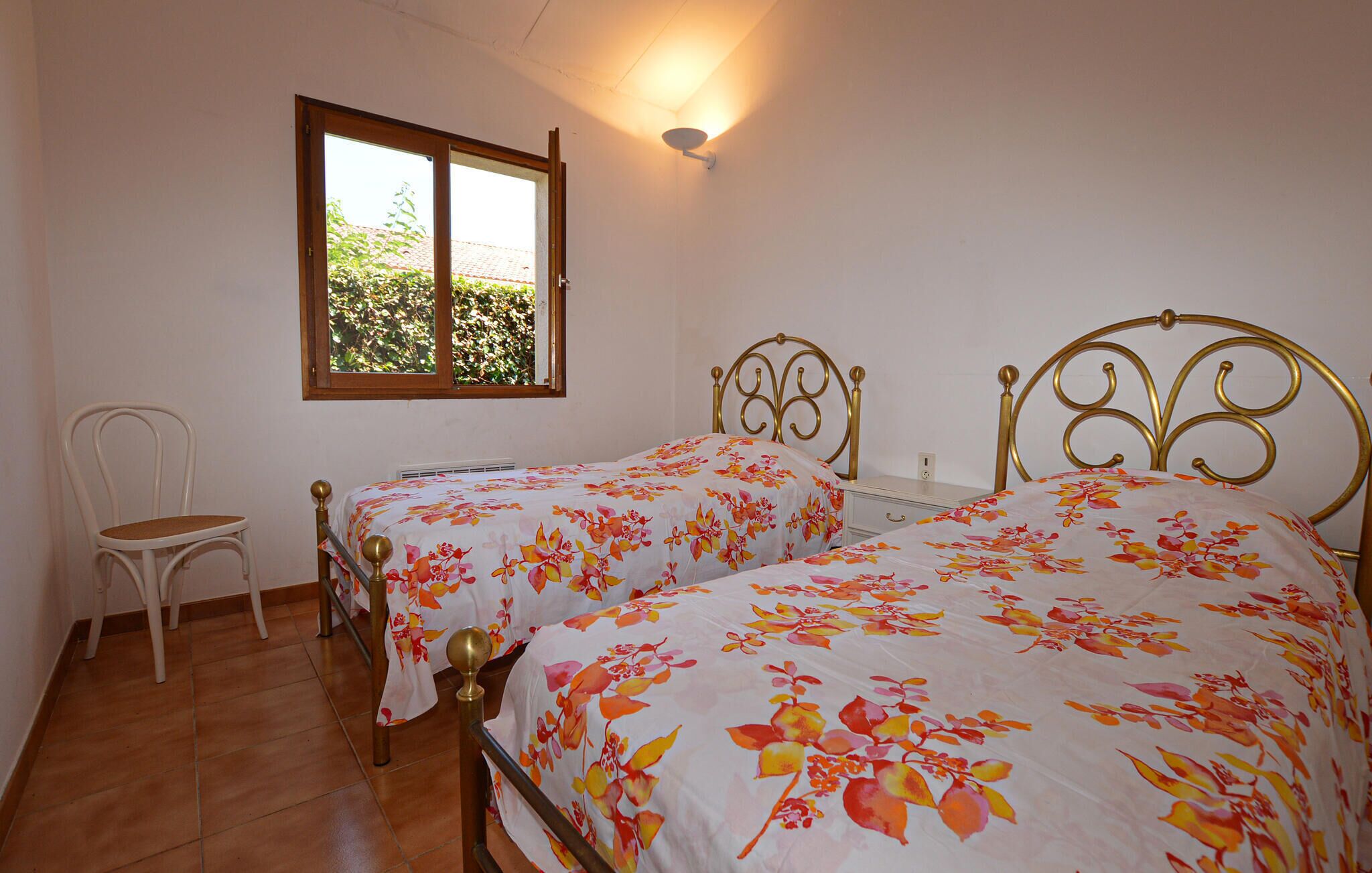 A bedroom with two beds, a chair, a window, and a wall-mounted light fixture.