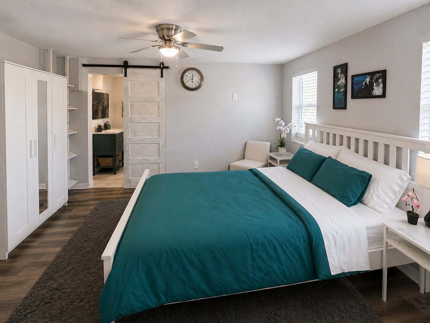 A bedroom with a bed, a nightstand, a chair, a ceiling fan, a clock, and a door leading to a bathroom.