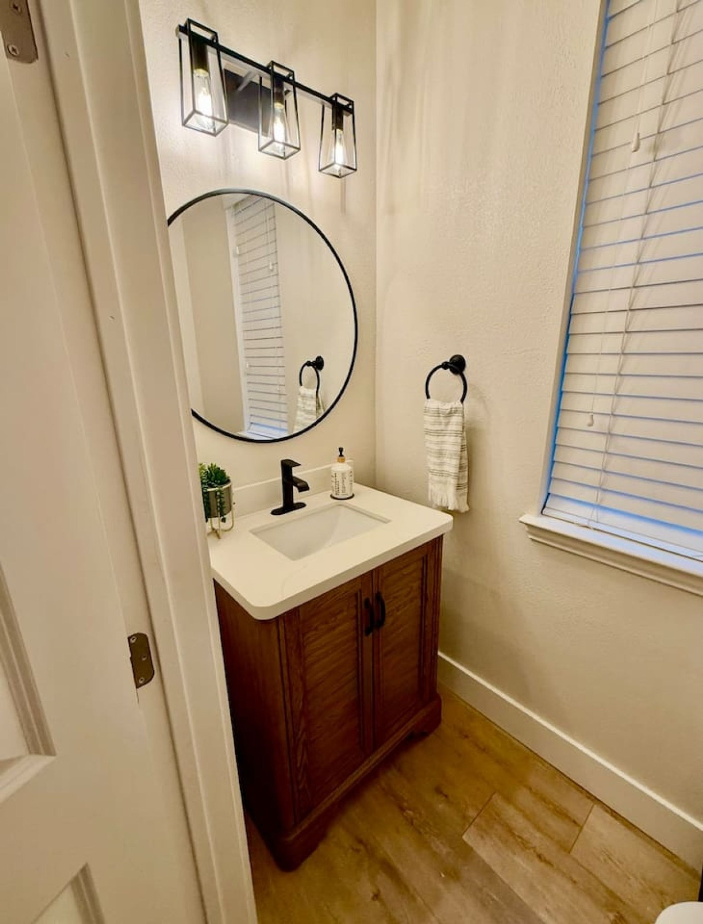 A bathroom with a wooden vanity, a round mirror, a sink, and a window with blinds.