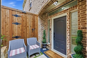 A brick house with a dark door, two wicker chairs with cushions, a small potted plant, and a welcome mat. - DFW Retreat w Pool 5 Min to Airport Sleeps 6 (Irving)