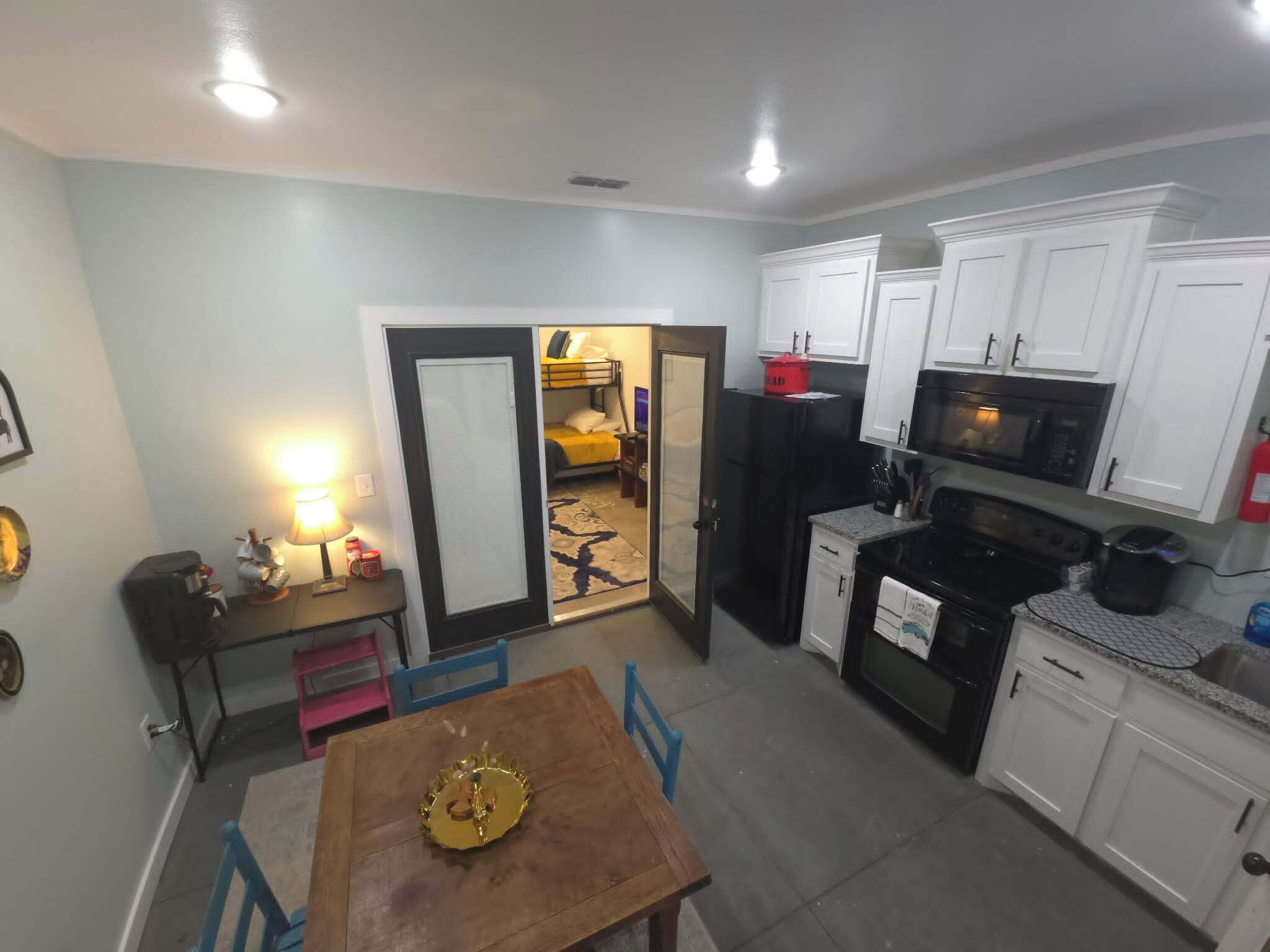 A compact kitchen with white cabinets, a microwave, a stove, and a refrigerator. There is a dining table with chairs and a small table with a lamp and candles.