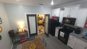 A compact kitchen with white cabinets, a microwave, a stove, and a refrigerator. There is a dining table with chairs and a small table with a lamp and candles.