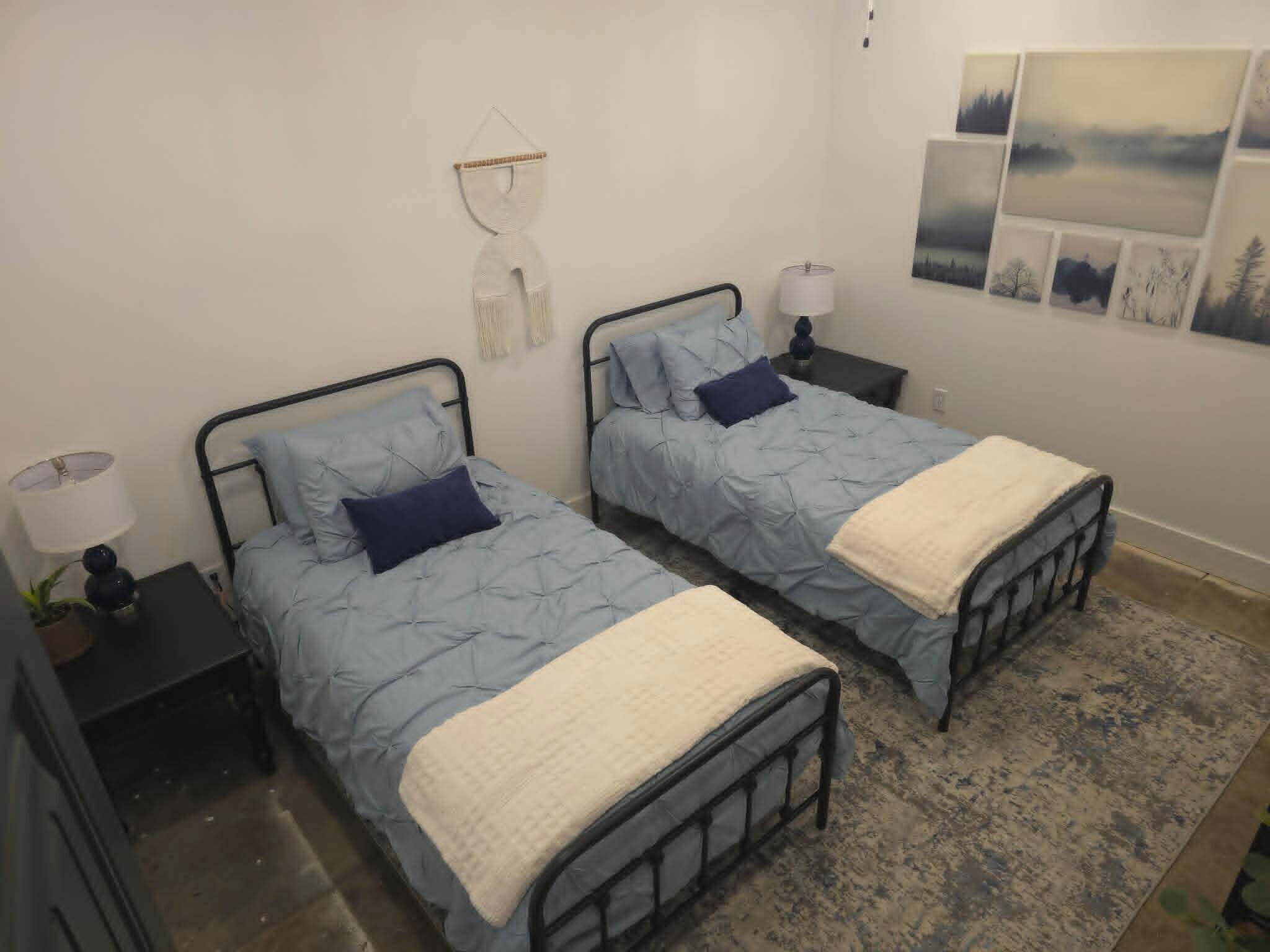 A bedroom with two beds, each with a blue quilt and a white throw blanket. There are bedside tables with lamps and a wall-mounted macrame decoration.