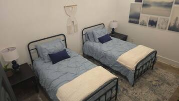 A bedroom with two beds, each with a blue quilt and a white throw blanket. There are bedside tables with lamps and a wall-mounted macrame decoration.