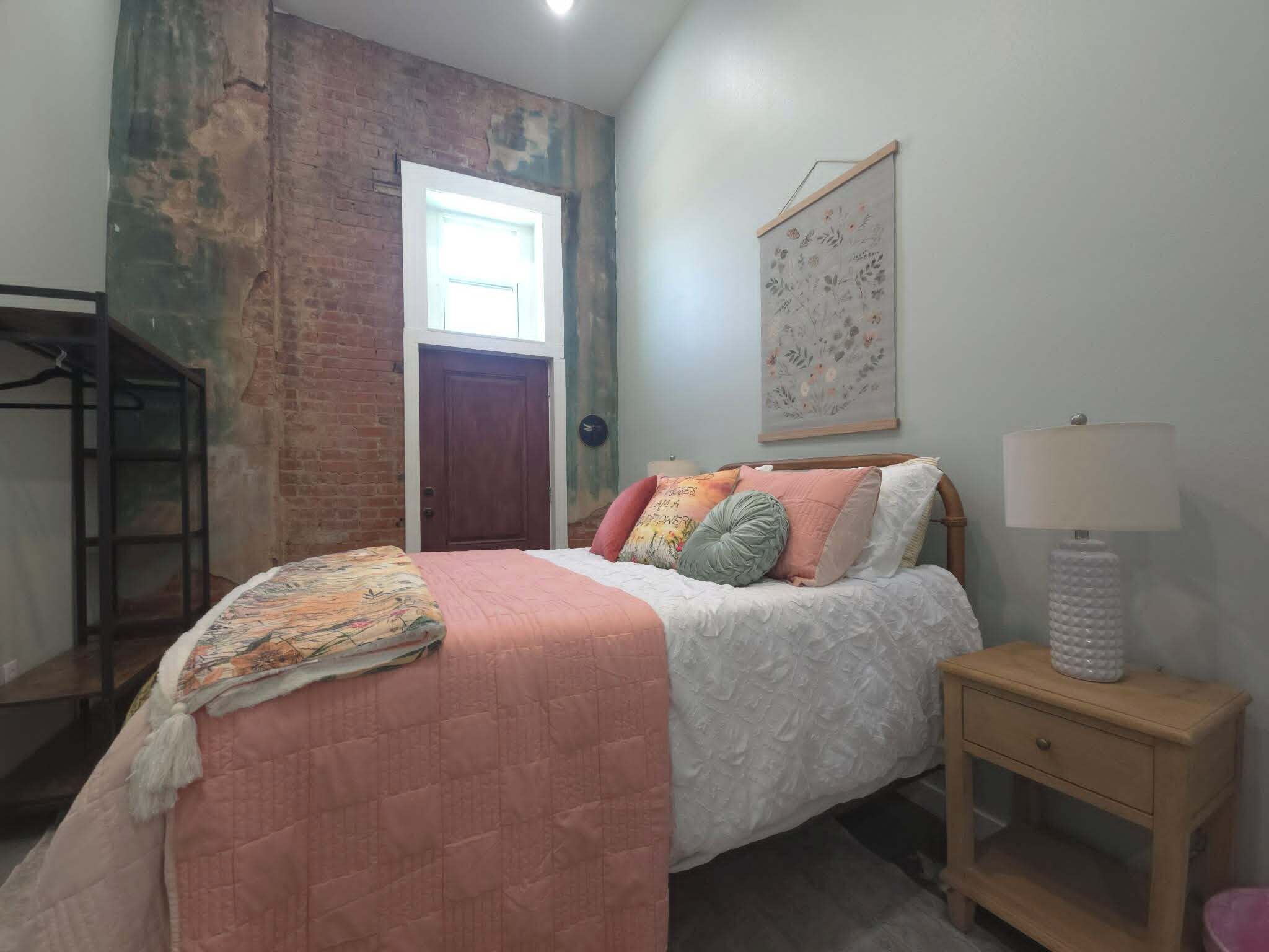 A bedroom with a bed, a nightstand, a lamp, a door, and a wall with a floral painting.