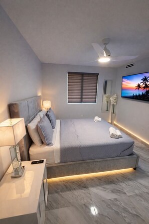 Ein modernes Schlafzimmer mit Bett, Nachttisch, Lampe, Fernseher und einem Fenster mit Jalousien.