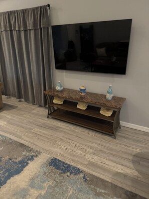 Un salon avec un téléviseur à écran plat fixé au mur, une console en bois agrémentée d’objets décoratifs, un rideau gris et un tapis à motifs.