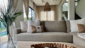 Una sala moderna con un sofá color beige, un florero de vidrio con plantas y una cesta tejida con mandos a distancia.