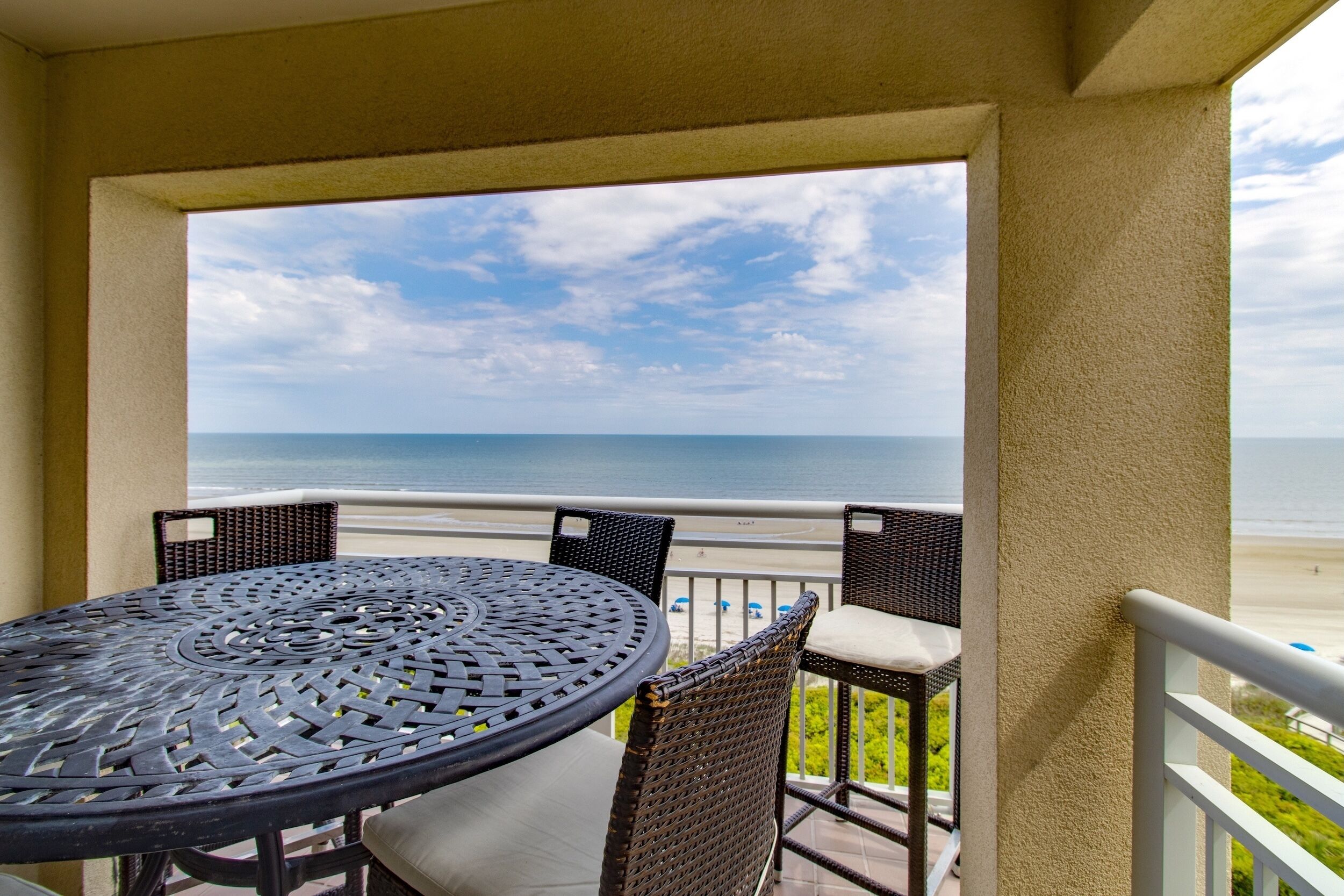 A balcony with a round table and wicker chairs, overlooking a beach.