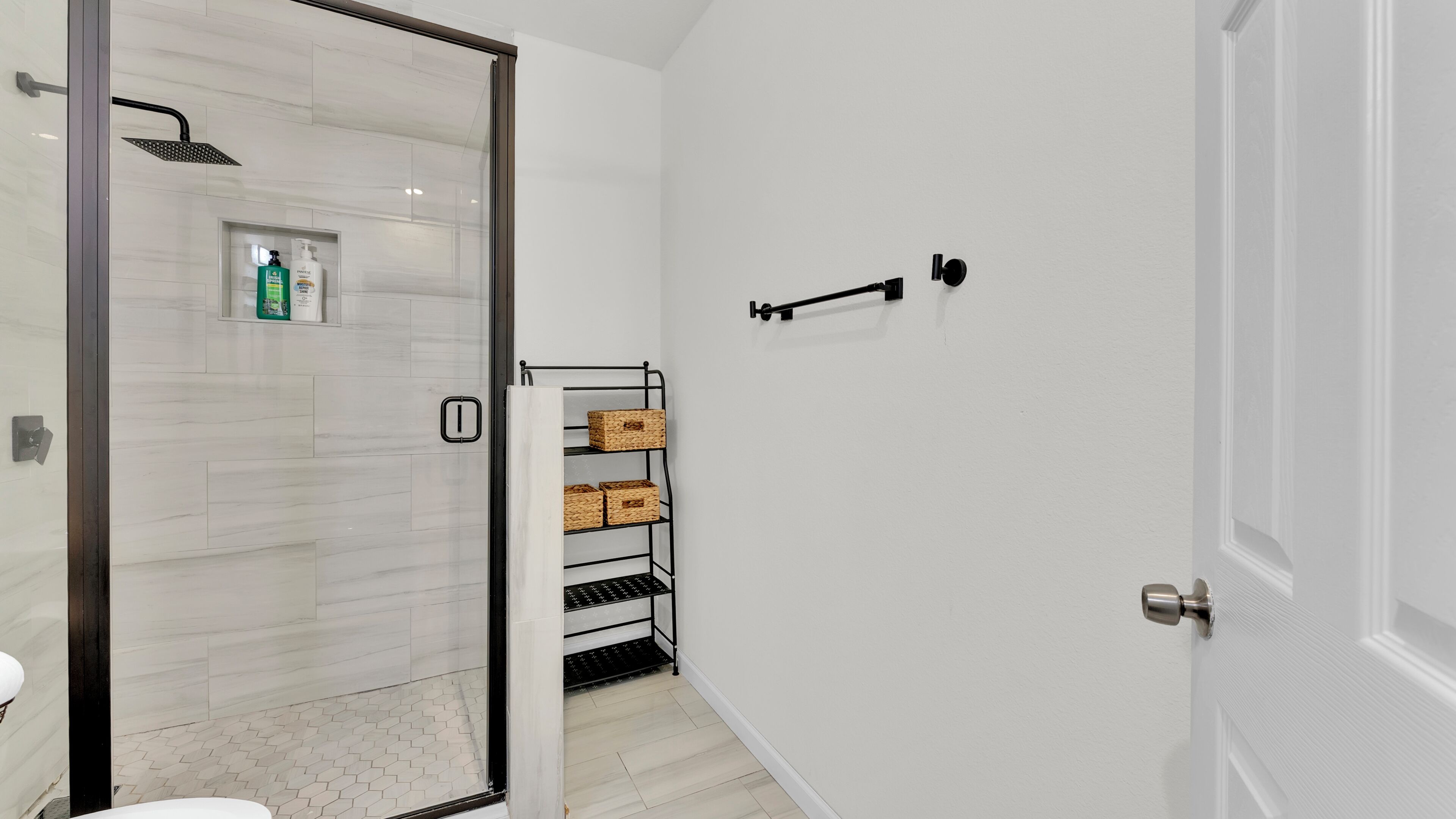 A modern bathroom with a glass shower enclosure, a white tiled floor, a white door, and a metal shelf with baskets.