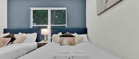 Una camera d'albergo con due letti, una finestra con tende, un comodino con lampada e un quadro appeso al muro.