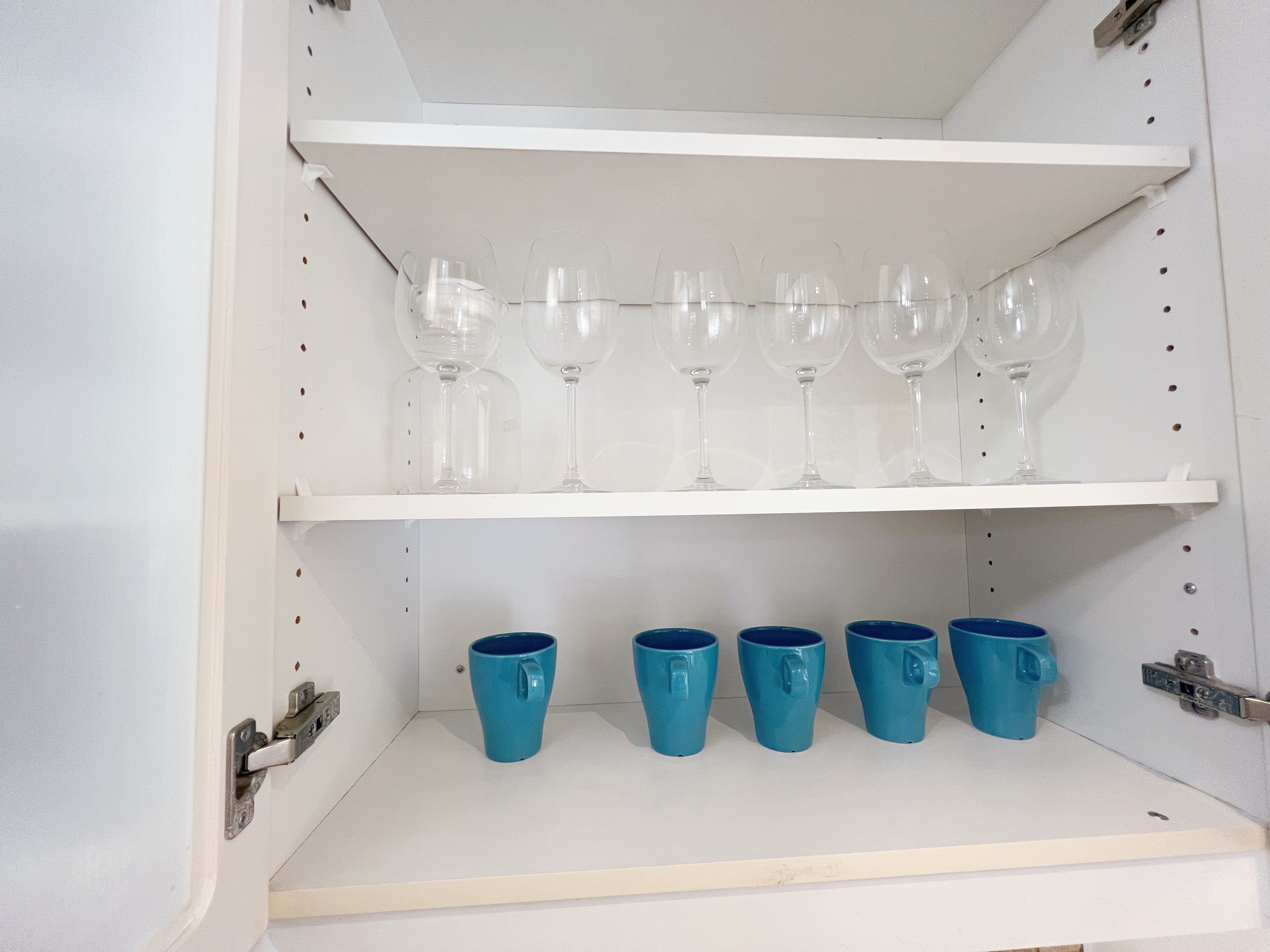 Een witte kast met planken waarop glazen wijn en blauwe mokken staan.