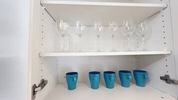 Een witte kast met planken waarop glazen wijn en blauwe mokken staan.