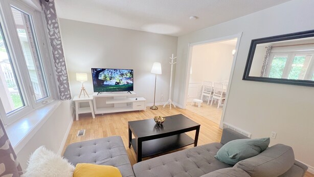 Ett vardagsrum med en grå soffa, ett svart soffbord, en platt-TV på ett vitt TV-bord, en lampa och en matplats med vita stolar.