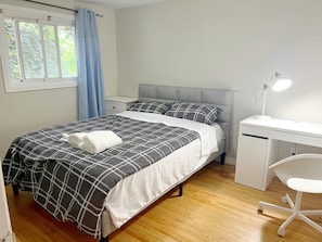 Una cama bien hecha con colcha a cuadros gris y blanca, una mesita de noche blanca, un cabecero tapizado en gris, un escritorio blanco con una silla y una ventana con cortinas azules.
