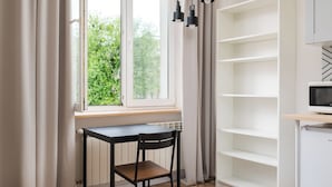 Ein Zimmer mit Fenster, Tisch, Stuhl, einer weißen Regaleinheit und einem Fernseher.