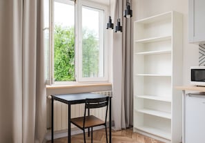 Ein Zimmer mit Fenster, Tisch, Stuhl, einer weißen Regaleinheit und einem Fernseher.