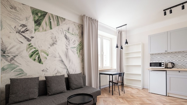 Ein modernes Wohnzimmer mit einer Couch, einem kleinen runden Tisch, einem Esstisch mit Stühlen und einer Küchenzeile mit Schränken und Geräten.